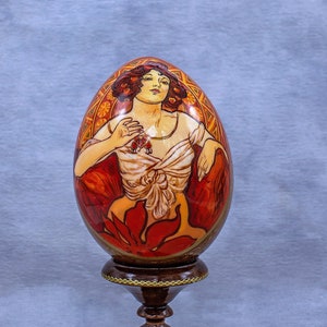 Könnte beinhalten: Ein handbemaltes Holzei mit einem detaillierten Bild einer Frau in einem roten und weißen Kleid. Das Ei wird auf einem dunkelbraunen Holzständer präsentiert.