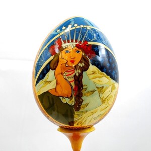 May include: A hand-painted wooden egg with a depiction of a woman wearing a crown and traditional clothing. The egg is on a wooden stand.