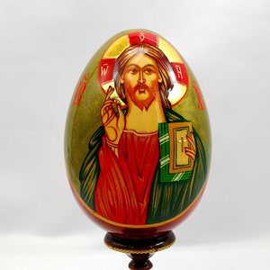 May include: A hand-painted wooden egg depicting Jesus Christ with a golden halo and holding a book. The egg is on a wooden stand with a brown base and gold trim.