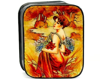 May include: A black wooden box with a painted image of a woman in a red dress holding a bouquet of flowers. The background is a landscape with trees and a stream. The box is decorated with gold accents.