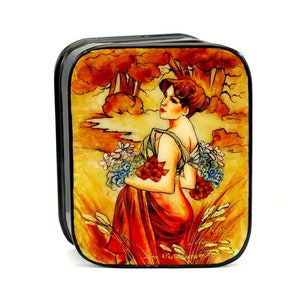 Op de afbeelding: Een zwarte houten doos met een geschilderd beeld van een vrouw in een rode jurk die een boeket bloemen vasthoudt. De achtergrond is een landschap met bomen en een beek. De doos is versierd met gouden accenten.