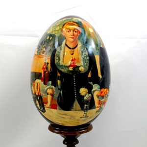 Pode incluir: Um ovo de madeira pintado à mão que retrata uma mulher com um vestido preto e um colar de renda branca, segurando uma taça de vinho e um prato de frutas. O ovo está em um suporte de madeira escura.
