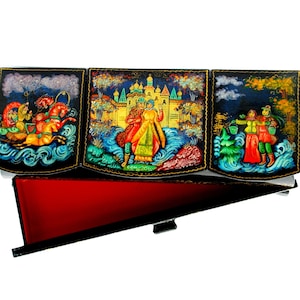 Op de afbeelding: Een zwarte houten doos met drie scharnierende panelen, elk beschilderd met een kleurrijke scène uit de Russische folklore. De doos is open en onthult een rood fluwelen interieur.