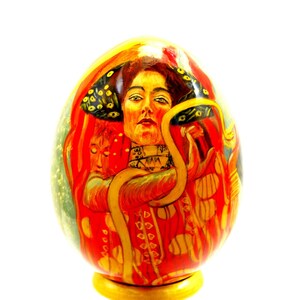 May include: A hand-painted wooden egg featuring a woman with a snake around her neck. The woman is wearing a red and gold dress with a peacock feather headdress. The egg is on a gold stand.