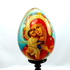 Op de afbeelding: Een handbeschilderd houten ei met een afbeelding van de Maagd Maria en het kindje Jezus. Het ei is versierd met gouden accenten en bloemmotieven. Het wordt gepresenteerd op een zwarte houten standaard.