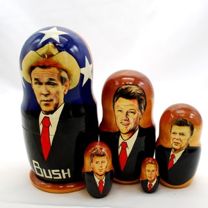May include: Set of five wooden nesting dolls, or matryoshka dolls, featuring portraits of American presidents. The largest doll depicts George W. Bush wearing a cowboy hat, with the inscription "Bush" on the base. The other dolls depict Bill Clinton, Ronald Reagan, and two smaller dolls with unclear faces.