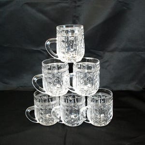 May include: Six clear glass beer mugs with handles are stacked in a pyramid formation. The mugs have a textured, patterned design. The background is black.