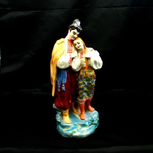 May include: A porcelain figurine of a couple in traditional Ukrainian clothing. The man is wearing a yellow and red embroidered shirt and a blue sash. The woman is wearing a white embroidered shirt with a yellow and blue patterned skirt. They are standing on a blue and white base.
