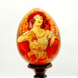 Könnte beinhalten: Ein handbemaltes Holzei mit einem detaillierten Bild einer Frau in einem roten Kleid vor einem floralen Hintergrund. Das Ei steht auf einem Holzständer.