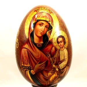Könnte beinhalten: Ein handbemaltes Holzei mit einer Darstellung der Jungfrau Maria und dem Jesuskind. Das Ei steht auf einem schwarzen Holzständer mit goldenen Akzenten.