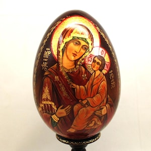 Op de afbeelding: Een handbeschilderd houten ei met een afbeelding van de Maagd Maria en het kindje Jezus. Het ei is versierd met bladgoud en ingewikkelde details. Het wordt gepresenteerd op een zwarte houten standaard.