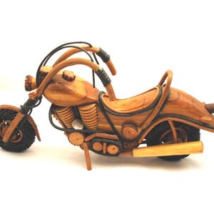 May include: A wooden motorcycle model with a brown finish. The model features black tires, handlebars, and seat detailing. The design includes visible engine components and a kickstand, showcasing a detailed craft.