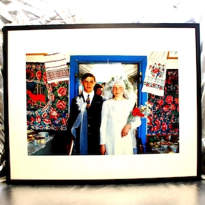 May include: A framed photograph depicting a wedding couple standing in a doorway. The person on the left wears a dark suit and tie, while the person on the right wears a white dress and veil. The background features vibrant floral textiles.