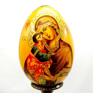 May include: A hand-painted wooden egg with a gold background and a depiction of the Virgin Mary and baby Jesus. The egg is on a wooden stand.