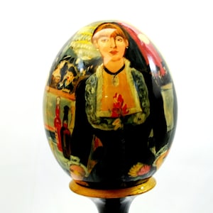 Op de afbeelding: Een decoratief ei met een geschilderd beeld van een vrouw in een zwarte jurk, die een rode bloem vasthoudt. Het ei heeft een glanzende afwerking en is gemonteerd op een zwarte standaard met een gouden voet. Het kunstwerk lijkt een schilderij te zijn.