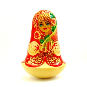May include: A traditional Russian nesting doll, or matryoshka doll, painted with a colourful design of a woman in a red dress with gold accents and flowers. The doll is made of wood and has a rounded shape.