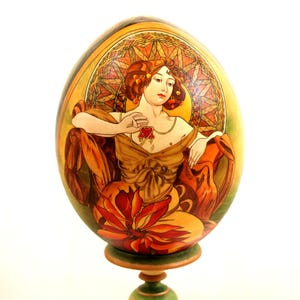 May include: A decorative wooden egg with an Art Nouveau design. The egg features a woman in a flowing gown, a red heart pendant, and a stained-glass style background. The egg is mounted on a green and brown wooden stand.