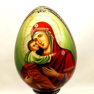 Op de afbeelding: Een handbeschilderd houten ei met een afbeelding van de Maagd Maria die baby Jezus vasthoudt. Het ei staat op een zwarte houten standaard met gouden accenten.