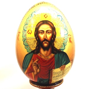 Può includere: Un uovo decorativo con un'icona dipinta a mano di Gesù Cristo con lunghi capelli castani e barba, che indossa una veste rossa e verde acqua. La figura è su uno sfondo dorato con un'aureola, tenendo in mano una pergamena con testo in cirillico.