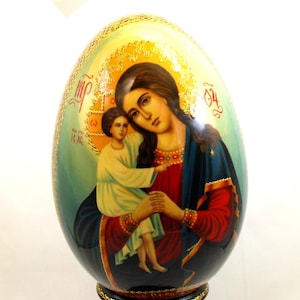 May include: A hand-painted wooden egg depicting the Virgin Mary holding baby Jesus. The egg is decorated with gold leaf and stands on a wooden base.