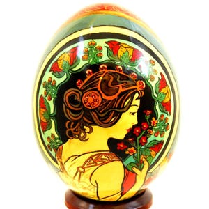 May include: A decorative egg featuring a painted illustration of a person in profile, surrounded by a circular border of red and yellow flowers. The person has dark hair styled with a red accessory and is holding a small cluster of red flowers.