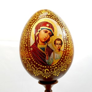 Könnte beinhalten: Ein hölzernes eiförmiges Ornament mit einem gemalten Bild der Jungfrau Maria und des Jesuskindes. Das Ei ist mit Blattgold und filigranen floralen Mustern verziert.