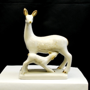 May include: A white porcelain figurine of a doe nursing her fawn. The doe has gold accents on her hooves and ears. The fawn is also white with gold accents on its hooves and ears. The figurine is on a white base.