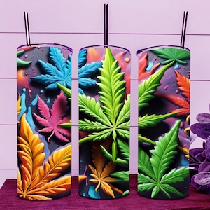 May include: Three stainless steel tumblers with colorful cannabis leaf designs. The leaves are in shades of yellow, pink, blue, and green, with a dark background and a splash of color. Each tumbler has a black straw.