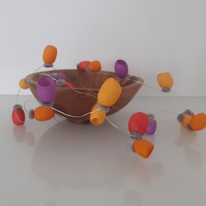 May include: A wooden bowl filled with a string of felt ornaments in orange, purple, and gray. The ornaments are strung together with a thin wire.