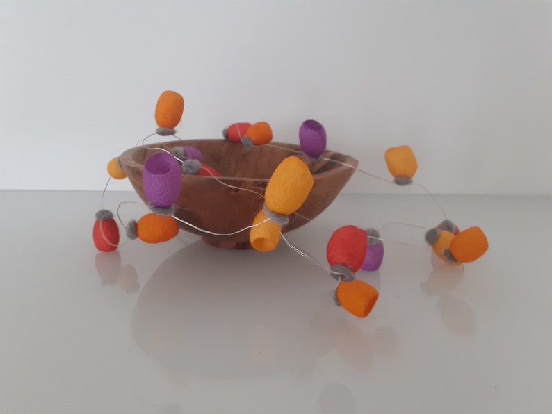 May include: A wooden bowl filled with a string of felt ornaments in orange, purple, and gray. The ornaments are strung together with a thin wire.