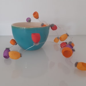 May include: A turquoise ceramic bowl with a string of felt balls in orange, purple, and red colors. The balls are strung on a thin wire and hang over the edge of the bowl.