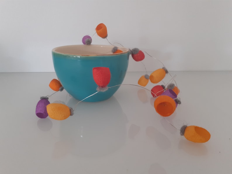May include: A turquoise ceramic bowl with a string of felt balls in orange, purple, and red colors. The balls are strung on a thin wire and hang over the edge of the bowl.