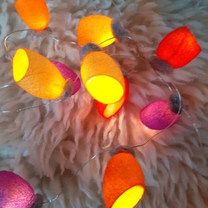 May include: A string of lights with felt lampshades in shades of orange, yellow, pink, and red. The lights are strung on a wire and are lit, casting a warm glow.