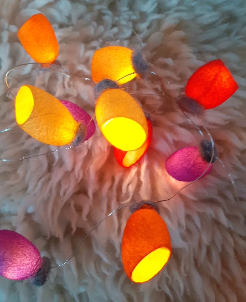 May include: A string of lights with felt lampshades in shades of orange, yellow, pink, and red. The lights are strung on a wire and are lit, casting a warm glow.