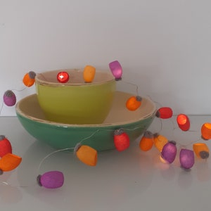 May include: A string of fairy lights with colorful felt covers in orange, purple, and red. The lights are wrapped around a green bowl and a yellow bowl.