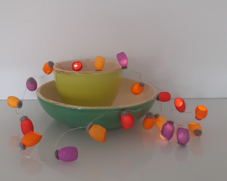 May include: A string of fairy lights with colorful felt covers in orange, purple, and red. The lights are wrapped around a green bowl and a yellow bowl.