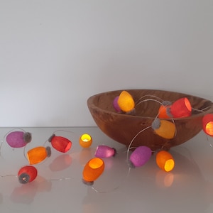 Könnte beinhalten: Eine Lichterkette mit Filzbezügen in Orange, Rosa und Rot. Die Lichter sind um eine Holzschale gespannt.