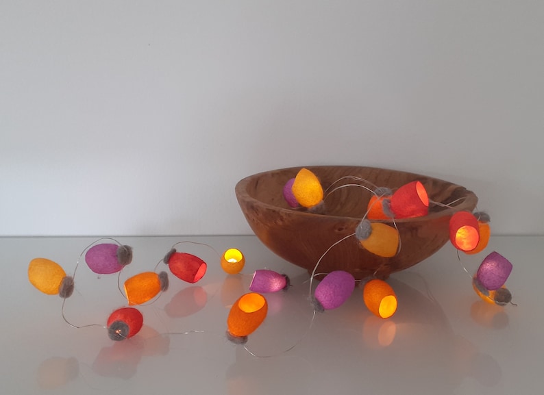 May include: A string of fairy lights with felt covers in orange, pink, and red. The lights are strung around a wooden bowl.