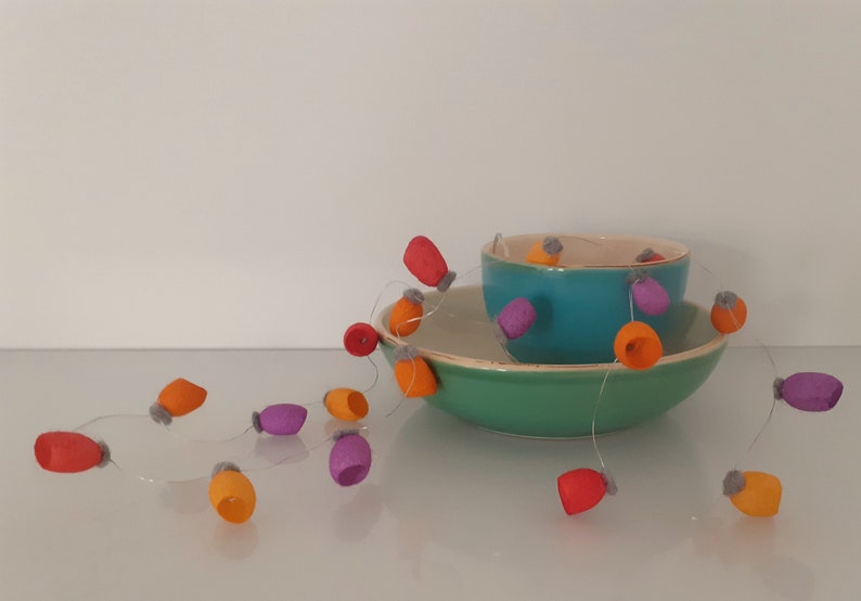 May include: A string of felt ornaments in orange, red, purple, and gray hangs between two bowls. The bowls are teal and green.