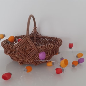 May include: A woven wicker basket with a handle sits on a white surface. A string of felt ornaments in orange, red, purple, and gray hangs from the basket.