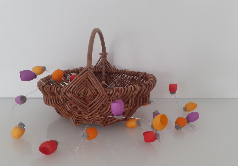 May include: A woven wicker basket with a handle sits on a white surface. A string of felt ornaments in orange, red, purple, and gray hangs from the basket.