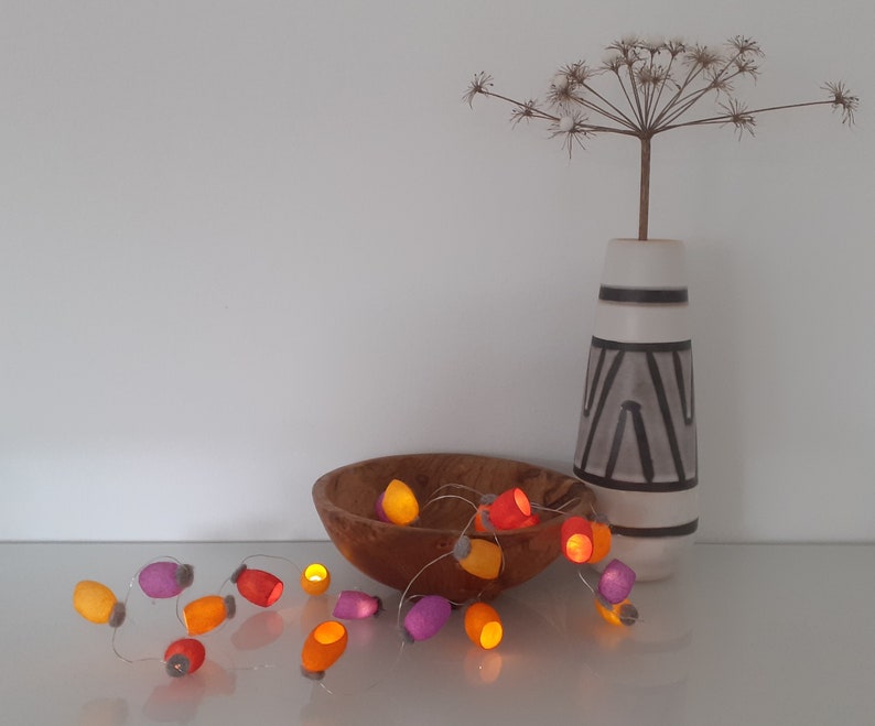 May include: A string of fairy lights with colorful felt covers in shades of orange, yellow, and purple. The lights are strung around a wooden bowl and are lit, casting a warm glow.