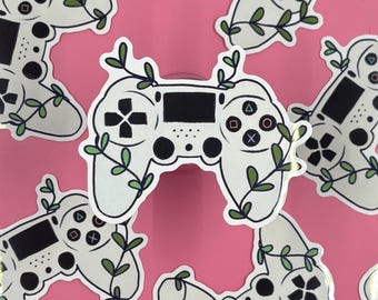 Puede incluir: Una pegatina de mando de videojuego blanco con detalles de botones negros e ilustraciones de enredaderas verdes con hojas pequeñas envueltas alrededor del cuerpo del mando, sobre un fondo rosa sólido con múltiples pegatinas repetidas.