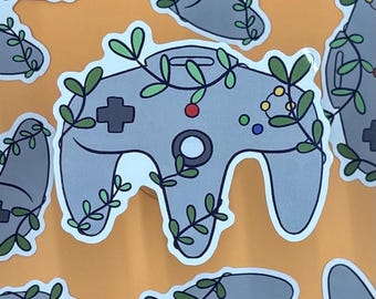 Puede incluir: Una pegatina troquelada que muestra un mando de videojuegos gris envuelto en enredaderas verdes con hojas pequeñas. El diseño del mando incluye una cruceta gris oscuro, un botón rojo y un grupo de cuatro botones pequeños de colores.