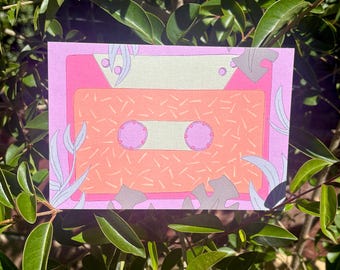 Puede incluir: Una impresión rectangular que presenta una ilustración estilizada de un casete. El casete tiene un borde rosa, una sección superior de color beige claro y un área central naranja con un patrón de pequeñas rayas blancas y dos carretes rosas circulares.