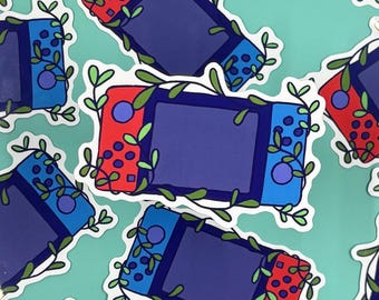 Puede incluir: Una colección de pegatinas troqueladas que muestran una consola de videojuegos portátil con un mando izquierdo rojo y uno derecho azul. Cada consola está ilustrada con enredaderas y hojas verdes que envuelven la pantalla y los botones.