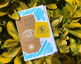 Puede incluir: Una pequeña libreta de papel marrón con las palabras "I can't sleep" escritas en la portada. La libreta tiene un diseño a rayas azul y blanco en la portada y una pegatina amarilla con un ojo dibujado a mano.