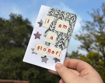 Puede incluir: Un diario hecho a mano con el texto "I am a stoner" en letras rojas. El diario tiene una cubierta blanca con estrellas decorativas y un diseño de hojas estampadas. El diario se sostiene contra un fondo de cielo azul y follaje verde.