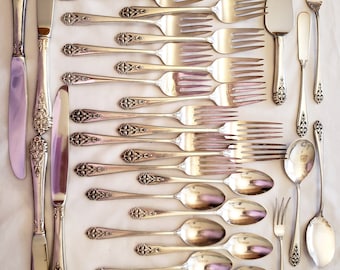 May include: An assortment of silver-colored flatware, including knives, forks, spoons, and serving utensils, arranged on a white surface. The handles feature an ornate floral design. The set includes various sizes of forks, spoons, and knives.