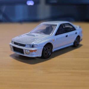 May include: A silver die-cast model car with black wheels. The miniature vehicle has a detailed front grille, headlights, and side mirrors. The car is a sedan style and sits on a wooden surface.